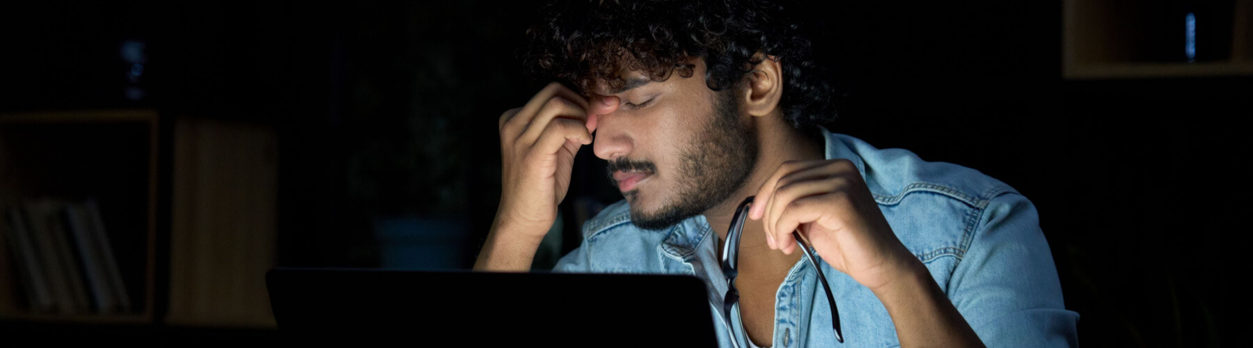 man lit by laptop screen holds his glasses and rubs his eyes