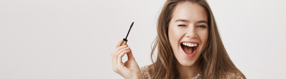 Indoor shot of positive attractive caucasian woman winking and smiling broadly while holding mascara and applying it