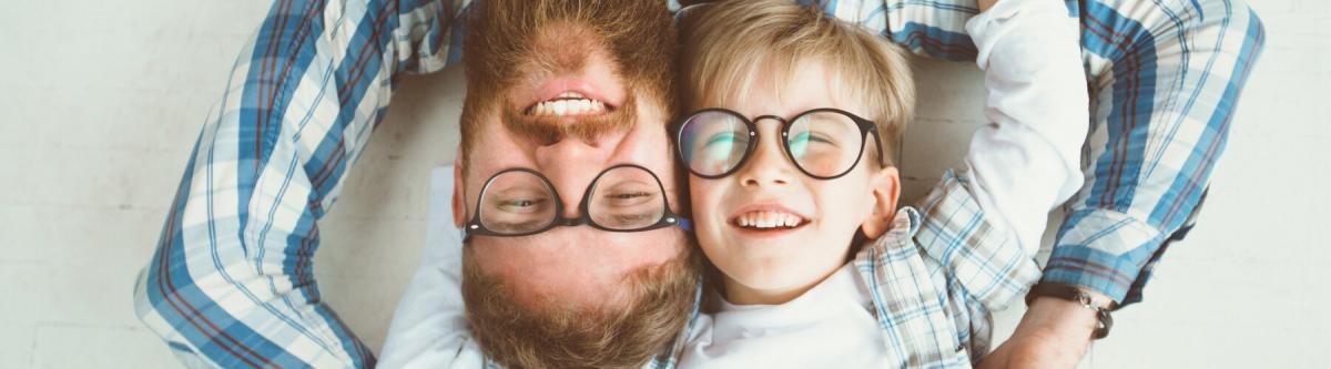 Top view of cute little boy and his handsome young beard dad, both in eyeglasses, smiling while lying with hands behind head on white floor.