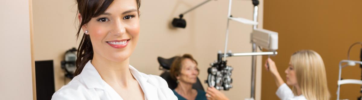 close up of a female optometrist smiling, in the background another optometrist is giving a woman an eye exam